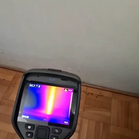 Cámara termográfica de J&R frente a un muro: en la pantalla se ve una franja clara vertical, una cañería de agua caliente que pasa por dentro